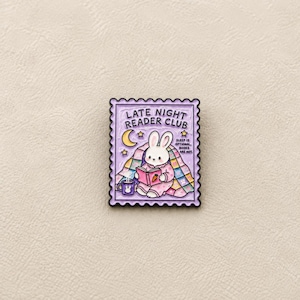 May include: A rectangular enamel pin shaped like a postage stamp with a scalloped edge. It features a white rabbit reading a book while wrapped in a colourful patchwork quilt. The text reads LATE NIGHT READER CLUB and SLEEP IS OPTIONAL, BOOKS ARE NOT.