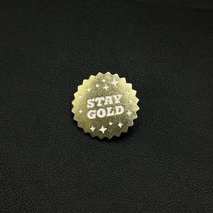 Peut inclure: Une épingle dorée en forme d'éclat d'étoile avec les mots "STAY GOLD" en lettres blanches. L'épingle a une surface texturée et est ornée de petites étoiles et de diamants.