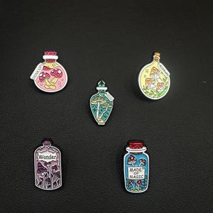 Peut inclure: Cinq épingles en émail représentant des champignons à l'intérieur de flacons de potion. Les épingles comportent des textes tels que "Drink Up", "Mushrooms", "Wonder" et "Made of Magic". Les flacons sont de différentes couleurs, dont le rose, le vert, le jaune, le violet et le bleu.