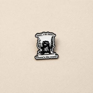 May include: A black and white enamel pin featuring a furry creature holding a coffee cup and a sword. The text above the creature reads "okay, i'm here..." and the text below reads "where's the work?" The design has a black border and white background.