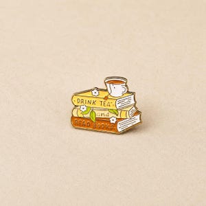 May include: A gold-toned enamel pin shaped like a stack of three books with a white teacup resting on top. The books feature yellow and orange covers with the text DRINK TEA and READ BOOKS printed in black, accented by small white flowers and green leaves.