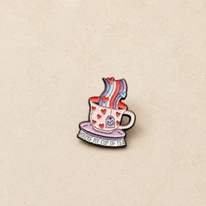 May include: A metal enamel pin shaped like a teacup on a saucer, decorated with red hearts. A colourful striped steam rises from the cup, which features a tea bag tag. The base of the saucer displays the black text, YOU'RE MY CUP OF TEA.