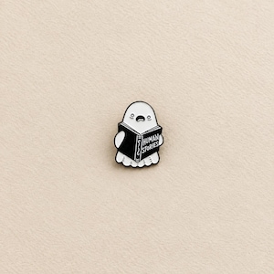 Peut inclure: Une broche en émail blanc en forme de fantôme avec une expression surprise, tenant un livre noir ouvert. Le livre porte le texte blanc 'HUMAN STORIES' sur sa couverture. La broche a un contour noir et un design plat et simple aux lignes épurées.