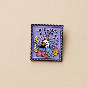 May include: A rectangular enamel pin shaped like a postage stamp with a light purple border. It features a white duck wearing a floral blanket, reading a pink book with a flashlight. Text reads LATE NIGHT READER CLUB and SLEEP IS OPTIONAL BOOKS ARE NOT.