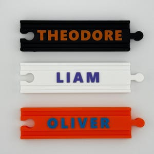 May include: Three colorful name tags in a train track design. The top tag is black with "THEODORE" in orange letters. The middle tag is white with "LIAM" in blue. The bottom tag is orange with "OLIVER" in blue.
