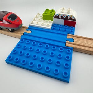 Puede incluir: Un juego de trenes de juguete con un tren rojo y blanco, un puente de plástico azul, vías de madera y bloques de construcción coloridos. El juego incluye un bloque verde, bloques blancos y una estructura arqueada roja y blanca.