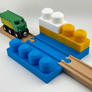 Könnte beinhalten: Ein grüner und gelber Spielzeugzug auf Holzschienen, der sich einer blau, gelb und weiß gehaltenen Blockstruktur nähert. Die Struktur hat blaue Zylinder und gelbe und weiße zylindrische Formen.