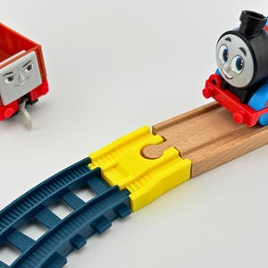 May include: A toy train set featuring a blue and red Thomas the Tank Engine train on wooden tracks, a red cargo car, and blue plastic tracks. The train is smiling. The set is on a white surface.