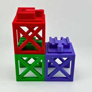 Könnte beinhalten: Drei farbenfrohe, geometrische Bausteine in Rot, Grün und Lila. Die Blöcke sind würfelförmig mit einem offenen, gitterartigen Design. Der rote Block sitzt auf dem grünen Block, und der lila Block ist daneben.