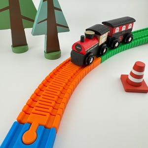 Puede incluir: Un juego de tren de juguete de madera con una locomotora roja y negra y un vagón negro sobre una vía naranja y verde. El juego incluye árboles de madera y un cono de tráfico a rayas rojas y blancas. La locomotora tiene el texto "100e" y "Le Toy Van".