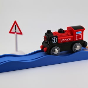 May include: A red and black toy train with the number 1 and the brand name "Hape" is on a blue track. A white road sign with a red triangle and exclamation point is in the background. The train is on a wooden track.