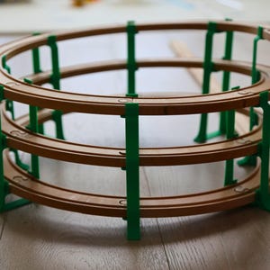 May include: A wooden toy train track set with a spiral design. The track is brown, supported by green vertical supports. The track is designed to allow toy trains to travel up and down the spiral.