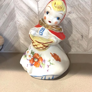 May include: A ceramic figurine of a young girl in a red hooded cape, holding a basket. The figurine has a white dress with floral details in orange, purple, and white. The girl has blonde hair and blue eyes.