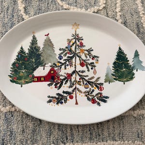 May include: An oval white ceramic serving platter featuring a festive holiday illustration. The design includes a central decorated Christmas tree, a small red house with a snow-covered roof, and several evergreen trees with ornaments.
