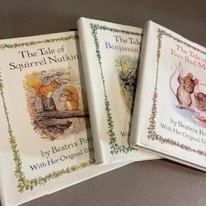 May include: Three vintage children's books by Beatrix Potter are displayed. The titles include "The Tale of Squirrel Nutkin", "The Tale of Benjamin Bunny", and "The Tale of Two Bad Mice". Each book has a cream-colored cover with illustrations and text.