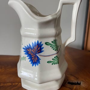 May include: Cream-colored ceramic pitcher with a blue and orange floral design. The pitcher has a unique octagonal shape with a handle and spout. Green leaves and stems accent the floral pattern. The pitcher is likely for serving beverages.