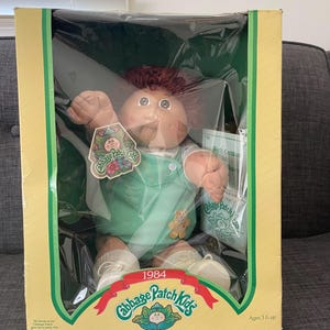 May include: A vintage Cabbage Patch Kids doll in its original box. The doll has brown hair, a peach-colored face, and is wearing a green outfit. The box is yellow with green trim and features the Cabbage Patch Kids logo and the text "1984".