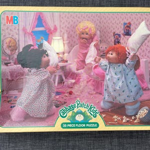 May include: A Cabbage Patch Kids 35-piece floor puzzle. The puzzle features a bedroom scene with three Cabbage Patch Kids dolls in pajamas, holding pillows. The puzzle has a pink and white color scheme.