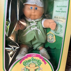 May include: A 1985 Cabbage Patch Kids Preemie doll with blue eyes, wearing a white cap, a green and white checked waistcoat, and matching shorts. The doll is inside its original packaging, which includes a birth certificate and adoption papers.