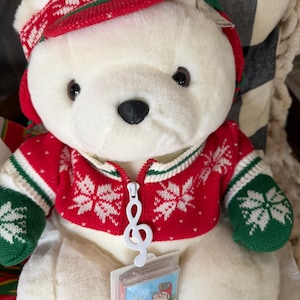 May include: A white plush teddy bear wearing a red and white knit cap and a matching red, white, and green snowflake-patterned zip-up sweater. The zipper pull is shaped like a white musical treble clef, and a small rectangular tag is attached.