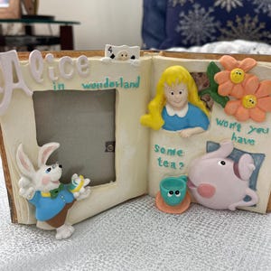 May include: A decorative Alice in Wonderland themed picture frame. The frame features illustrations of Alice, the White Rabbit, and other characters. The frame is beige with colorful accents and text.