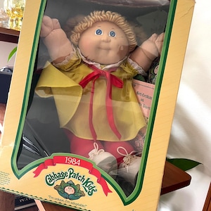 May include: A vintage Cabbage Patch Kids doll in its original box. The doll has blonde yarn hair, blue eyes, and is wearing a yellow dress with a red bow. The box is tan with green accents and the text "1984 Cabbage Patch Kids".