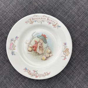 May include: White ceramic plate featuring a Benjamin Bunny illustration with a rabbit wearing a hat and carrying a red bag. The plate has the text "Benjamin Bunny" and "Beatrix Potter" in banners, with floral and bear accents.