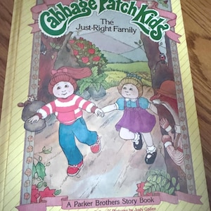 May include: A vintage Cabbage Patch Kids storybook titled "The Just-Right Family." The cover features illustrations of two Cabbage Patch Kids holding hands, set against a forest backdrop. The book is a Parker Brothers Story Book.