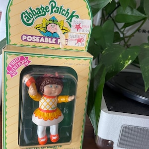 May include: A vintage Cabbage Patch Kids doll in its original packaging. The doll is wearing a yellow and white dress, white leggings, and orange shoes. The box reads "POSEABLE" and "FIRST EDITION". The doll's name is Lynnet Ellen.