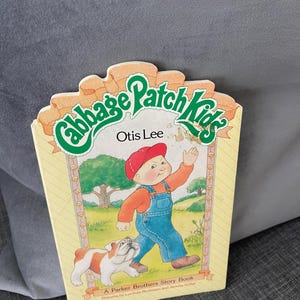 May include: A vintage Cabbage Patch Kids storybook titled "Otis Lee." The cover features an illustration of a young boy in overalls and a red cap, walking with a bulldog. The book has a yellow border with the Cabbage Patch Kids logo.