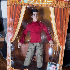 May include: An *NSYNC collectible marionette doll of JC Chasez in a red long-sleeve shirt and khaki pants. The doll is packaged in a box with the *NSYNC logo and the words "Collectible Marionette". The box also features a picture of JC Chasez.