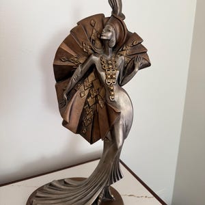 May include: A metallic bronze and silver-toned sculpture of a standing figure wearing a long, flowing gown with a large, pleated fan-like accessory behind the head and shoulders. The figure features a tall, pointed headpiece and a long necklace.