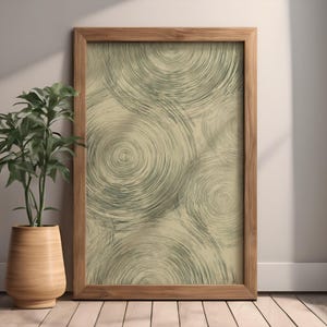 Könnte beinhalten: Ein gerahmter Kunstdruck mit einem abstrakten Muster aus sich überlappenden konzentrischen Kreisen in Grau- und Grüntönen auf beigem Hintergrund. Das Kunstwerk wird in einem Naturholzrahmen präsentiert, der an einer weißen Wand lehnt.
