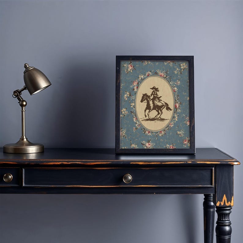 May include: A framed print featuring a sepia-toned illustration of a person riding a horse, set against a floral patterned background. The print is in a black frame and rests on a black desk with a brass lamp.