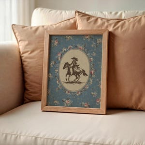 May include: A framed print featuring a vintage illustration of a person riding a horse. The artwork is set against a blue floral background with a light brown wooden frame. The image is resting on a white surface.