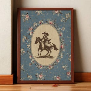 May include: Framed artwork featuring a sepia-toned illustration of a person riding a horse, set within an oval frame. The background is a blue floral pattern, and the frame is a dark brown color. The artwork is leaning against a white wall and a wooden floor.