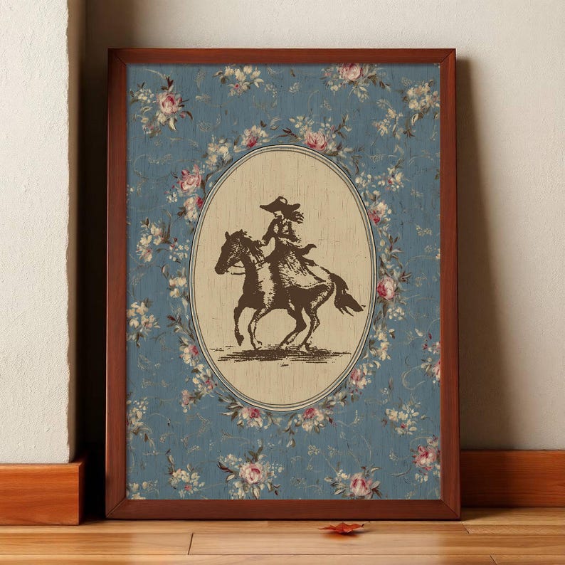 May include: Framed artwork featuring a sepia-toned illustration of a person riding a horse, set within an oval frame. The background is a blue floral pattern, and the frame is a dark brown color. The artwork is leaning against a white wall and a wooden floor.