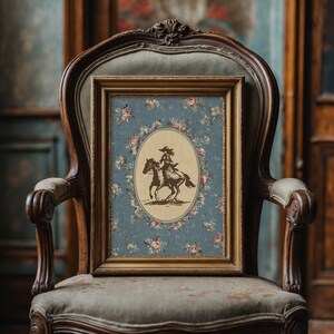 May include: A framed vintage print featuring a sepia-toned illustration of a person riding a horse. The print is set against a blue floral background and framed in gold. The artwork is displayed on a vintage upholstered chair.