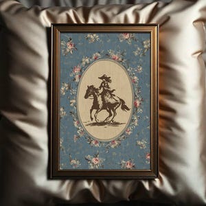 May include: Framed artwork featuring a sepia-toned illustration of a person riding a horse. The image is set within an oval frame, surrounded by a floral wreath on a blue background. The frame is a dark brown color.