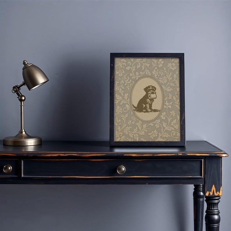 Puede incluir: Una impresi&oacute;n enmarcada con una ilustraci&oacute;n en tonos sepia de un perro sosteniendo un objeto, sobre un fondo floral. La impresi&oacute;n est&aacute; en un marco negro y se encuentra en un escritorio negro con una l&aacute;mpara de escritorio de lat&oacute;n.