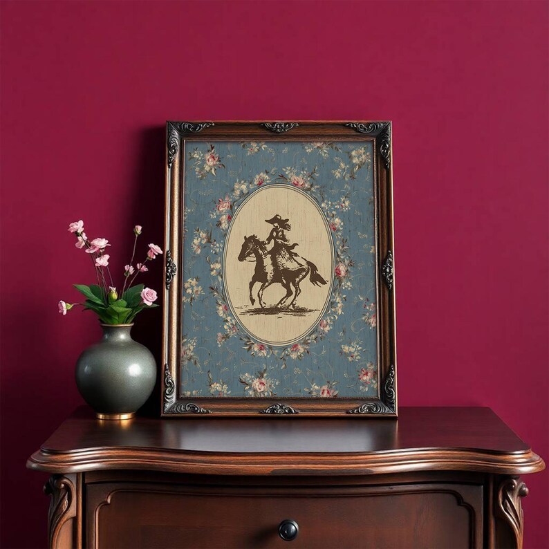 May include: A framed print featuring a sepia-toned image of a person riding a horse, set against a floral patterned background. The frame is dark brown with ornate detailing. The print is displayed on a wooden dresser.