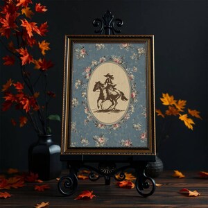 May include: A framed print featuring a sepia-toned illustration of a person riding a horse, set against a floral patterned background. The frame is gold, and the print is displayed on a black easel. The overall aesthetic is vintage and decorative.