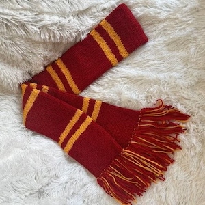 May include: A red and gold striped scarf with a fringe. The scarf is made of a soft, knitted fabric.