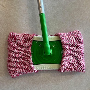 May include: A green and silver mop with a pink and white knitted cleaning pad. The mop has a handle and a round head with a white plastic ring around the edge.