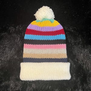 May include: A colorful striped knitted beanie with a white pom pom on top. The beanie has a white brim and is made with a variety of colors, including red, blue, yellow, pink, and black.