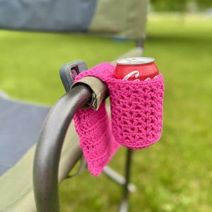 May include: A bright pink, crocheted drink holder attached to a folding chair armrest. A red Coca-Cola can sits inside the holder. The chair is silver and khaki, set against a green grassy background.