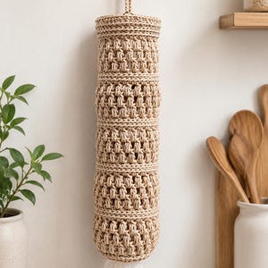 Op de afbeelding: Een beige, handgemaakte gehaakte zakjesdispenser hangt aan een witte muur. De cilindrische zakhouder is gemaakt van dik, getextureerd garen en heeft een lus om op te hangen. Een witte plastic zak is zichtbaar aan de onderkant. Keukendecoratie.