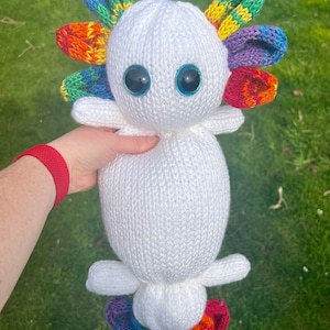 May include: A white knitted axolotl plush toy with rainbow colored fins and tail. The toy has large blue eyes.