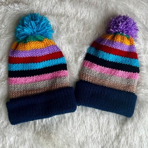 May include: Two striped knitted hats with pom-poms. The larger hat has a blue, yellow, red, pink, and black striped pattern. The smaller hat has a purple, yellow, red, pink, and black striped pattern. Both hats have a dark blue brim.