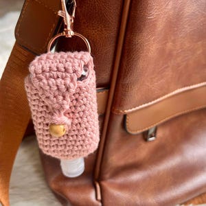 May include: A small, pink crocheted bottle holder with a wooden bead closure, attached to a rose gold clip. The holder is designed to carry a small bottle, and is shown hanging from a brown leather bag.